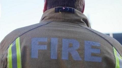 A shot of a firefighter taken from behind his shoulders. The top of his light brown jacket reads: "Fire". He is looking towards a grey sky.