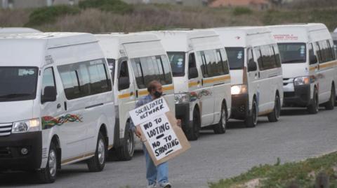 A taxi driver stands in front of parked taxi vans in Cape Town, holding a placard that reads “No to looting, no to shooting”