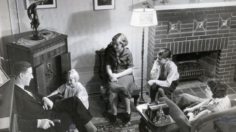 A black and white image of a family with three children gathered around a wooden wireless radio set in the left corner