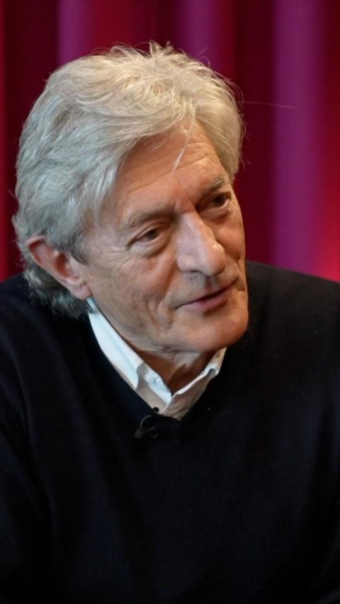 Nigel Havers in a black jumper and white shirt sitting in front of a purple backdrop.