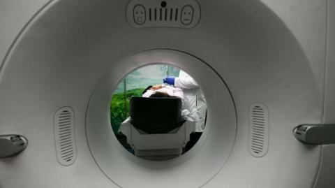 An unidentified woman is laid down with someone with a white coat attending to her. The image is shot through the central ring of a large circular white scanning device.
