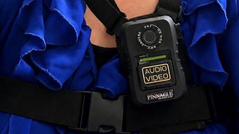 A black body camera with the words "audio" and "video" in large yellow lettering. The rectangular camera has a lens at the top and is being worn by a woman wearing a blue blouse.