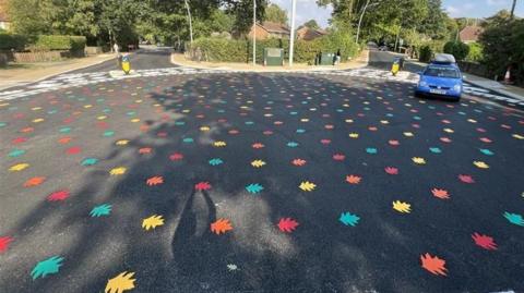 The middle of a roundabout painted with red, orange, green and yellow leaves. A car is entering the circle.