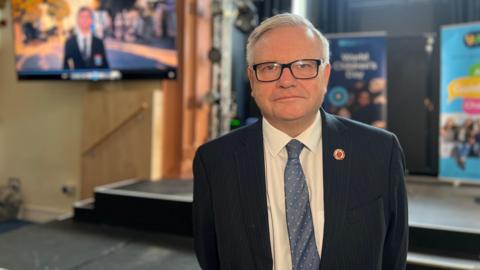 A man wearing a suit looking towards the camera. He is wearing glasses. 