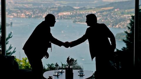 Two figures in silhouette shake hands with a lake visible behind them, at talks in Switzerland between the US and Iran