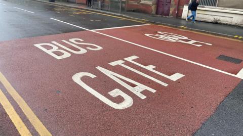 Street scene in Corporation Street, Preston. Showing a red brick building in the background. The road says 'bus gate' in white on a red background on the left hand side. A person is walking on the far side of the road.