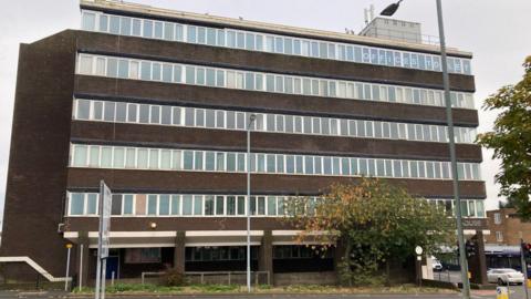 A large office block, six storeys high with lots of windows and bleak brown walls.