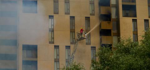 A firefighter on an aerial ladder sprays a hose reel onto a tower block whose balconies are blackened from heat and smoke. 