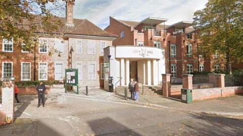 A view of Epsom and Ewell town hall - red brick buildings with tall windows in a leafy street. A few people are standing outside.