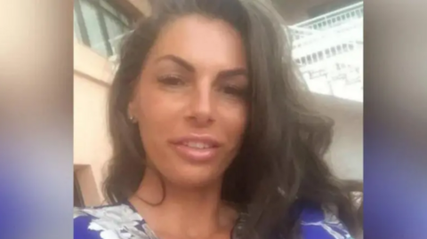 A woman with long brown hair smiling at the camera. She is wearing a blue and white top