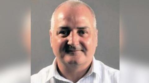 A head and shoulders shot of Sexton who is wearing an open neck white shirt. He is smiling and has very short grey hair. 