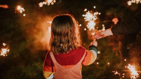 Child is seen holding a sparkler with an adult's hand also in shot helping her keep it upright. She is facing away from the camera standing on a grass surface.