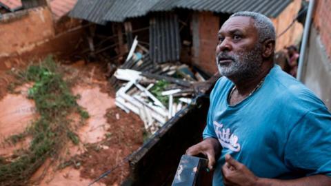 Isaque Alves da Silva, 58, talks from the balcony of a house affected by a landslide in Ipatinga. He is wearing a blue T-shirt. Below him, a house overflowing with mud can be made out. 