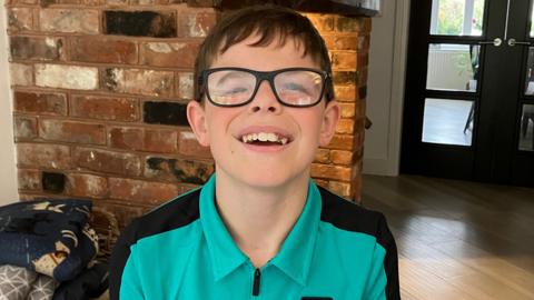 A boy with short black hair, glasses, a green and black shirt, smiles as he stands in a house. A brick wall is behind him as well as a door leading into another room.