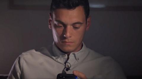 A man with brown hair and wearing a light blue shirt looks in contemplation at his phone