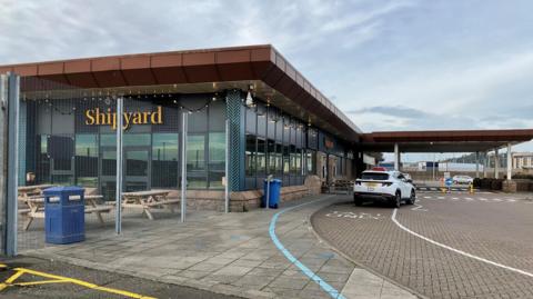 An outside photo of the Shipyard cafe at Elizabeth Harbour in Jersey. There is a white car parked outside. It's a large building with long glass windows. The word shipyard is prominent in yellow writing on the wall. There are benches and a bin behind a metal fence.