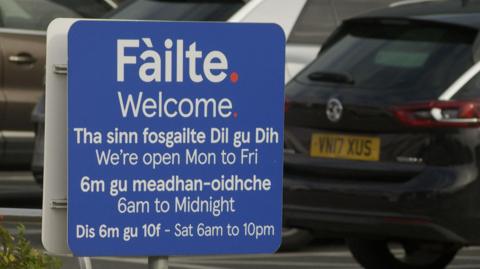 Welcome sign outside the Stornoway tesco, it lists the stores opening hours in English and Gaelic