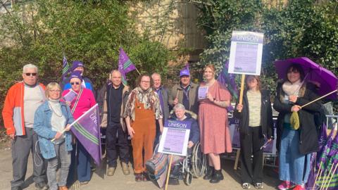 A group of protestors outside the full council meeting on Wednesday. They are holding purple Unison flags and placards. There are 12 people in attendance.