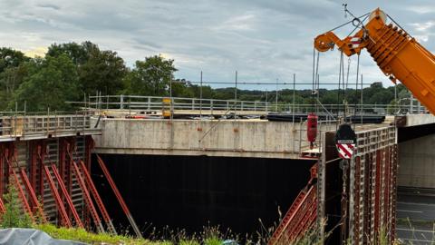 A large concrete bridge structure is under construction, supported by red steel formwork and scaffolding