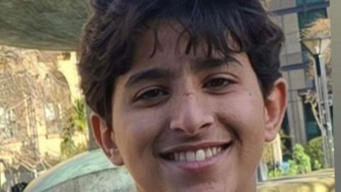 A boy smiles at the camera. He has brown hair, which falls over his forehead. The picture was taken outside, with several trees in the background.