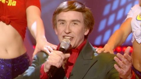 Steve Coogan as Alan Partridge with microphone and gesturing to crowd