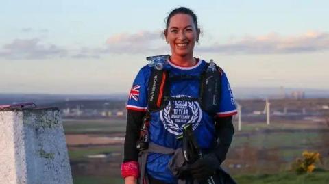 A woman wearing a blue shirt and a rucksack with attachments for water bottles is standing on top of a hill with a white-painted trig point next to her. In the distance are a series of wind turbines.