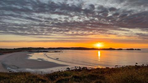 L’Ancresse in Guernsey. There is a large beach and the sun appears to be setting. The sky is orange and blue. The sky is cloudy.