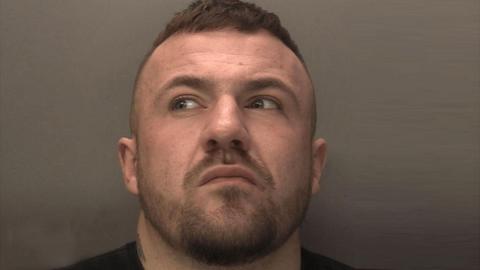 A police mugshot of a man with brown hair who is looking away from the camera.