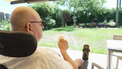 A man in a wheelchair sits eating an ice cream cone overlooking the garden of the care home 