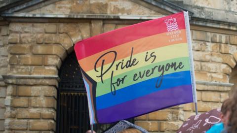 A rainbow flag is waved in front of a historical building in Oxford on 7.6.25. It has the words "Pride is for everyone" and the logo of Oxford City Council, which supported the event.