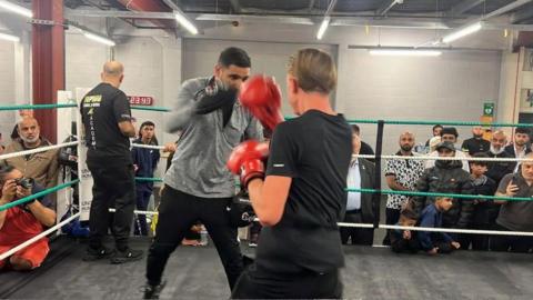 Khan, who is wearing a grey T-shirt and black trousers, is holding his hands up which have boxing gloves on. A person is standing in front of him with their back to the camera wearing red boxing gloves.