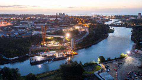 An aerial view of a river with a number of cranes lifting a section of the bridge. The city lights can be seen behind the river and the sea at the back.