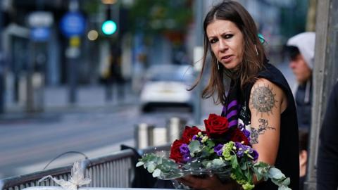 A woman with dark brown hair and black eyeliner has tattoos on her arm. She is standing on a pavement and is holding a bunch of flowers with red roses, purple flowers and green leaves.