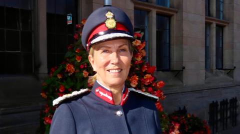 Elizabeth Barnes, a woman wearing a navy coloured ceremonial uniform with a red collar and white epaulettes. She is standing outdoors, in front of a stone building with a floral arrangement.