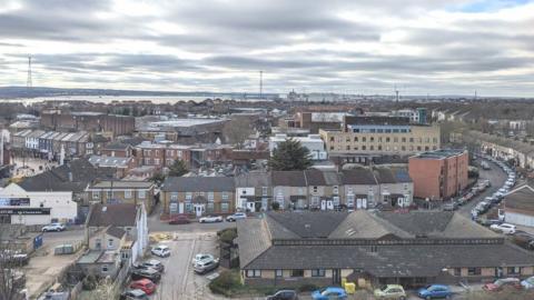 The rooftops of terraced houses and offices spread out near Grays town centre, with the River Thames and pylons visible in the background, set against a cloudy sky.