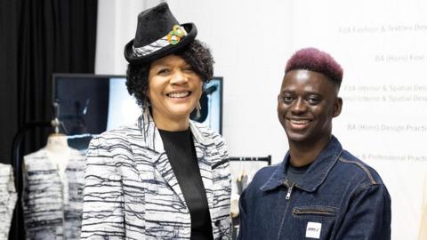 Chi Onwurah wearing a black and white, almost tiger-print suit jacket over a black top. She is wearing a small black hat with a strip of fabric and two orange and green circles. She is smiling into the camera and standing to the right of Lamin Gibba who has pink hair and is wearing a denim jacket. He is also smiling into the camera