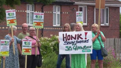 Five women and one man hold placards on the side of the road. One of the signs reads: Honk! Show support for S13 Greenbelt