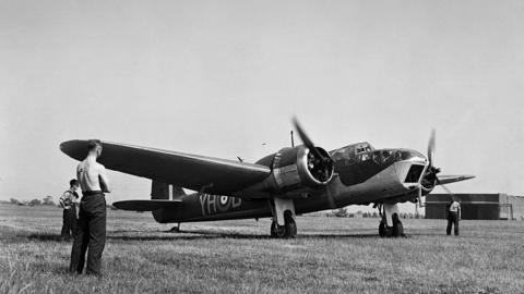 A Bristol Blenheim Mk IV, YH-B, (V6240) light bomber aircraft of No 21 squadron is warming up its engines before take-off from a grass airfield in 1940. Three men are looking on - the man nearest the camera is topless.