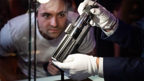 A man wearing white gloves holds the lightsaber while a man in a white t-shirt inspects it. It is a relatively small looking silver item, with four holes along the top and a narrow pole coming out of it. 