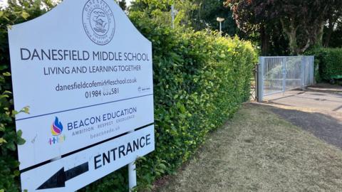 Image of the Danesfield Middle School sign. It is white, and has the logo  at the top. There is also a logo for Beacon Education. There is an arrow pointing left to the entrance at the bottom. 