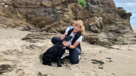 Sarah Walker smiling at the camera next to her black dog on a beach. She's knelt down and is stroking the dog.