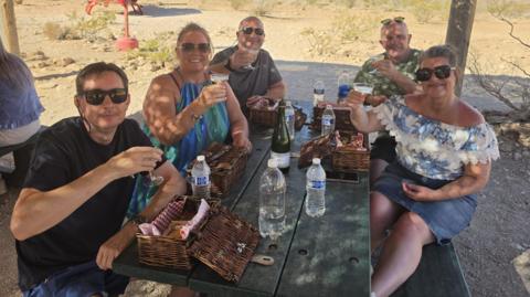 A group of five people sat at a picnic table in the Gran Canyon. They are all looking at the camera smiling and holding a drink aloft.