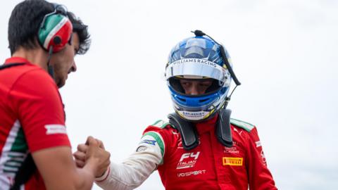 Bondarev dressed up in his racing kit and helmet shakes hands with a member of the racing team in Italy. It is a cloudy day.