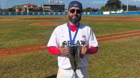 A man in dark glasses stands holding a silver trophy on a baseball pitch