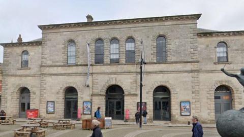 A view of the exterior of the theatre. It is an old sandy stone, two-story building. The front has five archways with doors. Seven arched windows are on the first floor. 