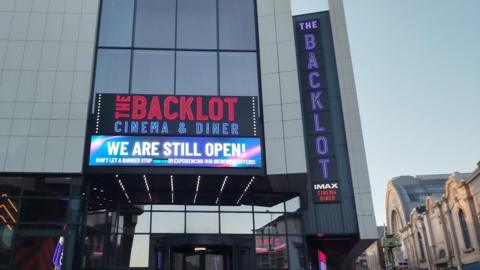 Front of the cinema which is a combination of glass and panels with the dome of Blackpool Winter Gardens to its right. Two signs spell out "The Backlot" cinema and diner in lights, while a screen above the entrance states in capital letters: "We are still open!"