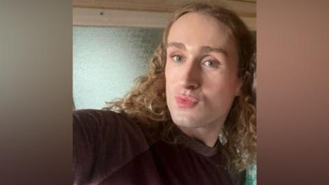 A person with long curly blonde hair and a black top looks at the camera.