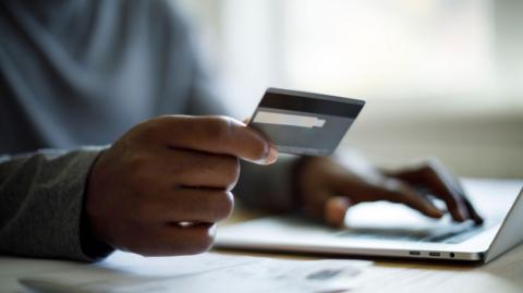 A man wearing a grey sweater sitting at a desk in front of a laptop holds a credit card in one hand between his index finger and his thumb, while his other hand is tapping the keys on the laptop. We cannot see the face of the man.