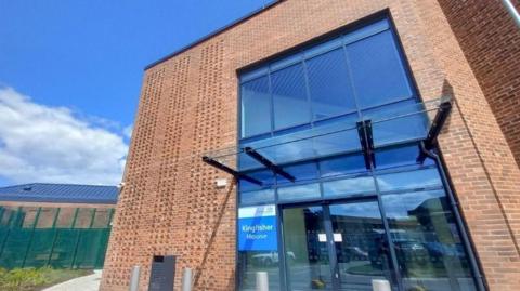 A new brick building with a large glass entrance, with green fencing in the background