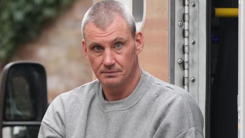 A man with short grey hair wearing a grey tracksuit top. He is being taken out of a prison van and staring at the camera.